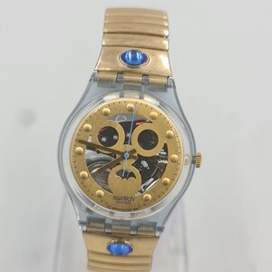 Puede incluir: Un reloj Swatch dorado con una caja de plástico transparente y una correa de metal dorado. La esfera del reloj es esqueletizada, revelando el funcionamiento interno del reloj. El reloj tiene piedras preciosas azules en la correa.