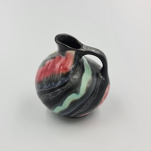 Puede incluir: Una pequeña jarra redonda de cerámica con asa. La jarra es principalmente negra con coloridos acentos arremolinados de rojo, rosa y verde. La abertura y el asa son negras. Es un objeto decorativo.