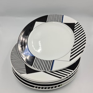 May include: A stack of white ceramic plates with a modern geometric design. Each plate features a black and white striped border, black triangles, and a thin blue accent line. The plates are stacked on a white surface.