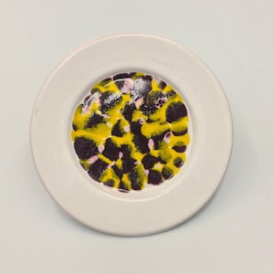 May include: A round, white ceramic plate with a speckled rim. The center of the plate features a vibrant design of yellow and dark purple splotches, with hints of pink. The plate is set against a plain white background.