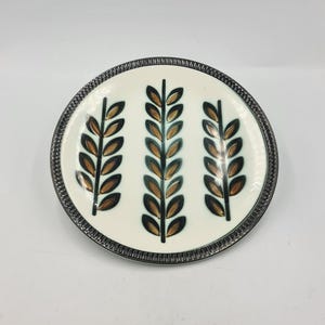 Può includere: Un piatto in ceramica bianca con un bordo argentato presenta un design stilizzato di tre spighe di grano marroni e verdi.