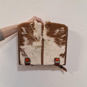May include: A rectangular bag with a brown and white cowhide pattern. The bag has a brown leather trim, two front buckles with orange accents, and a brown leather strap. The bag is held by a person's hand.