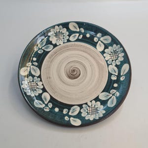Op de afbeelding: Een ronde keramische plaat met een teal rand en een wit en bruin spiraalvormig midden. De plaat is versierd met witte bloemen- en bladerpatronen rond de rand. De plaat heeft een diameter van ongeveer 25 cm.