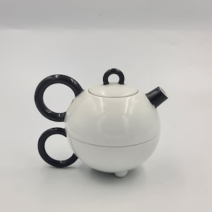 May include: A spherical white teapot featuring a glossy finish, a black circular handle, a black lid knob, and a black spout. The teapot is supported by small, rounded feet at the base, creating a distinct, balanced silhouette with high-contrast colors.