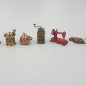 May include: A set of miniature kitchen and household items, including a mortar and pestle, a teapot, a coffee grinder, a sewing machine, a bundt cake pan, a frying pan, and an iron. The items are made of metal and are painted in various colors, including gold, copper, red, and black.
