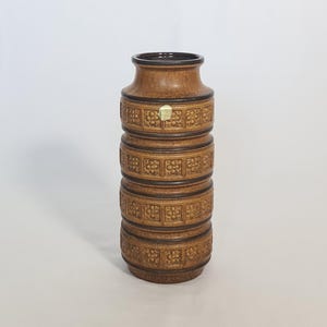 May include: A brown ceramic vase with a cylindrical shape. The vase features a series of horizontal bands with a floral pattern. The top and bottom rims are a darker brown. A small sticker is affixed to the front of the vase.