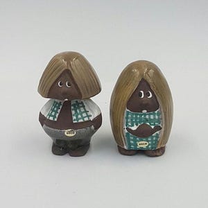 Puede incluir: Dos figuritas de cerámica de personas con cabello castaño y piel morena. Llevan camisas a cuadros verdes y blancas y tienen la palabra "Deco" en su ropa.