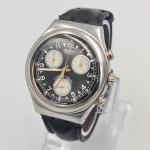 May include: A Swatch chronograph wristwatch featuring a round silver-tone metal case, a black dial with white Arabic numerals, three cream-colored sub-dials, gold-tone hands, and a black leather strap. The dial displays the text Swatch Swiss.