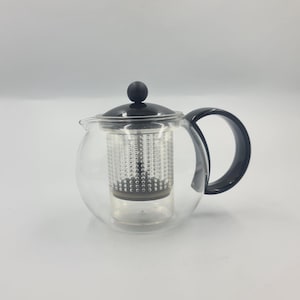 May include: A clear glass teapot with a black handle and lid. The teapot has a cylindrical infuser basket inside. The handle and lid are black. The teapot is designed for brewing tea.