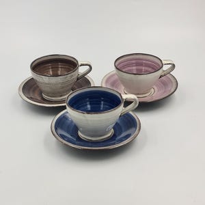 May include: Three ceramic tea cups and matching saucers arranged in a triangular formation. Each cup and saucer set features a distinct colour scheme: one brown, one pink, and one blue, all with a white glazed exterior and a handle.