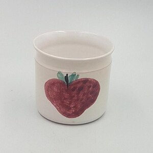 May include: White ceramic pot with a hand-painted red strawberry design. The strawberry has green leaves and a black stem.