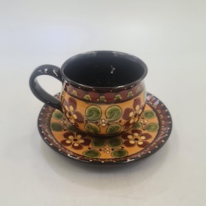 May include: A ceramic teacup and saucer set. The cup and saucer are decorated with a floral pattern in shades of brown, green, and yellow. The cup has a black handle and interior. The saucer has a matching design around the rim.