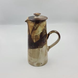 Op de afbeelding: Een hoge cilindrische keramische kan met deksel, voorzien van een gevlekt glazuur in tinten crème, beige en donkerbruin. De kan heeft een kleine schenktuit, een gebogen handvat aan de zijkant en een plat deksel met een kleine knop.