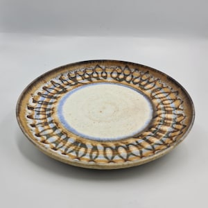 May include: A ceramic plate with a cream-colored center, a light blue ring, and a brown and tan patterned border. The plate has a slightly raised edge and a rustic, handmade appearance. The plate is likely for serving food.