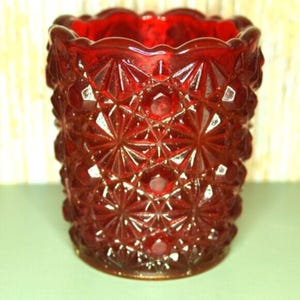 May include: A ruby red glass votive candle holder with a detailed geometric pattern. The holder has a scalloped rim and a textured surface, creating a vintage aesthetic. The candle holder is approximately 3 inches tall.