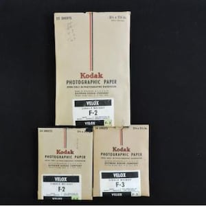 May include: Three packages of Kodak photographic paper, labeled "VELOX" and "F-2" or "F-3". The paper is in beige packaging with red and black vertical stripes. The packages are 8 x 10 inches.
