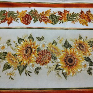 May include: A white tablecloth with a repeating pattern of sunflowers, leaves, and fall foliage. The tablecloth has a gold and orange stripe border.