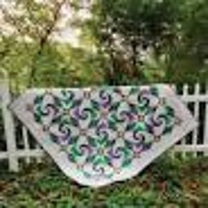 May include: A handmade quilt with a white background and a repeating pattern of green, purple, and teal swirls. The quilt is draped over a white picket fence in a garden setting.