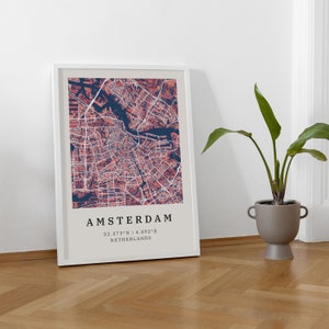 May include: A framed print of a map of Amsterdam, Netherlands. The map is in shades of red and blue, with the city's canals and streets clearly visible. The text "AMSTERDAM" is printed in black at the bottom of the map, along with the coordinates "52.3733&deg;N | 4.8922&deg;E".