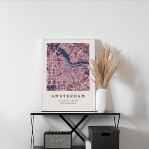 May include: A framed print of a map of Amsterdam, Netherlands. The map is in shades of pink and blue, with the city's canals and streets clearly visible. The text "AMSTERDAM 52.373&deg;N | 4.892&deg;E NETHERLANDS" is printed below the map.