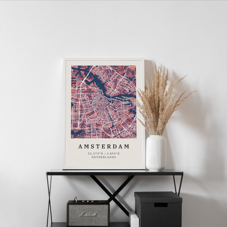 May include: A framed print of a map of Amsterdam, Netherlands. The map is in shades of pink and blue, with the city's canals and streets clearly visible. The text "AMSTERDAM 52.373&deg;N | 4.892&deg;E NETHERLANDS" is printed below the map.