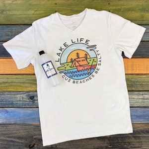 May include: White V-neck t-shirt with a circular graphic design featuring a lake scene with a tent, a canoe, and the text "LAKE LIFE CUZ BEACHES BE SALTY".