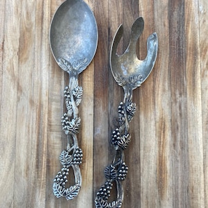 May include: Silver-toned serving spoon and fork set. The handles are adorned with a grape vine design, including leaves and clusters of grapes. The spoon has a large, oval bowl, and the fork has three tines. The set rests on a wooden surface.
