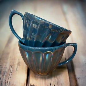 May include: Two teal-colored teacups are stacked on a wooden surface. The cups have a ridged design and curved handles. The cups have an iridescent sheen, reflecting colors of gold and green. The background is a blurred wooden surface.