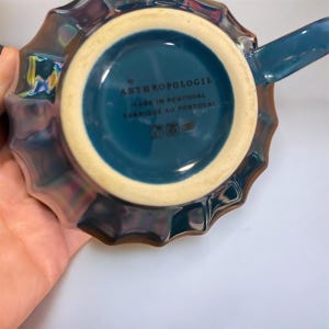 May include: A teal-colored ceramic mug with a handle, featuring a scalloped rim and iridescent finish. The bottom of the mug has the text "by ANTHROPOLOGIE MADE IN PORTUGAL FABRIQUE AU PORTUGAL". The mug is held by a hand.