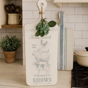 May include: A white wooden cutting board with a handle, featuring a farm-themed design with text that reads "Farm Fresh" and "Livestock Shows." The design includes illustrations of farm animals. A sprig of greenery is tied to the handle.