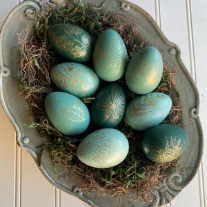 May include: A decorative gray ceramic bowl filled with moss and several teal-colored Easter eggs. The eggs are adorned with gold leaf designs, showcasing various botanical patterns. The bowl sits on a white surface.
