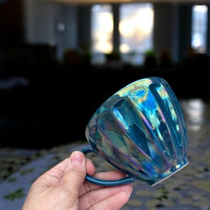 May include: A teal-colored ceramic mug with a unique, ridged design. The mug has an iridescent finish that reflects multiple colors. The handle is a matching teal color. The mug is designed for hot beverages.