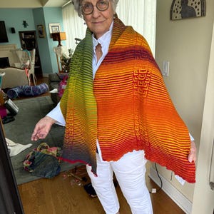 May include: A colorful, hand-knitted shawl with a gradient of colors from green to orange and red. The shawl features a geometric pattern and is worn over a white shirt and pants. The shawl is a unique, handmade accessory.