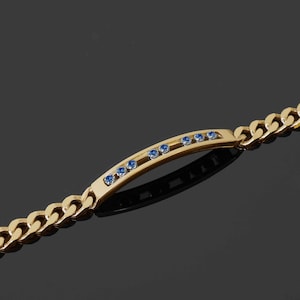 May include: A gold bracelet with a curb chain design. The bracelet has a rectangular bar in the centre, decorated with small, round, blue gemstones. The bracelet is shown against a dark background.