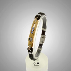 May include: A black leather bracelet with a gold and silver clasp. The clasp features a small ship's wheel design and blue gemstones.