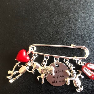 May include: Silver brooch with charms depicting characters and symbols from The Wizard of Oz. Charms include a red heart, a horse, a scarecrow, ruby slippers, and a disc with the phrase "there's no place like home."