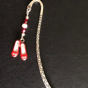 May include: A silver bookmark with a curved top and a decorative swirl design. A charm of red ballet slippers hangs from a beaded strand of red and white beads. The bookmark is on a black surface.