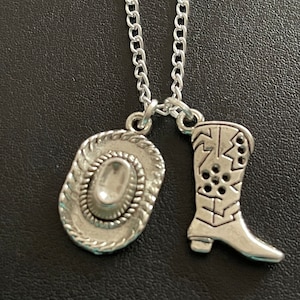 Puede incluir: Collar plateado con dos colgantes: un sombrero de vaquero y una bota de vaquero. El colgante del sombrero tiene un ala y una corona detalladas, mientras que el colgante de la bota muestra intrincados diseños recortados. La cadena es de estilo eslabón delicado.