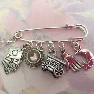 Peut inclure: Broche épingle de sûreté argentée avec plusieurs breloques. Les breloques comprennent des cartes à jouer, une tasse à thé, un train, une main et un croissant de lune avec une étoile. Une perle de cristal transparente est suspendue.