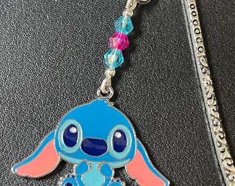 May include: A metal bookmark featuring a blue cartoon character with large ears and a light blue body. The bookmark has a decorative silver hook and a strand of blue and pink beads.