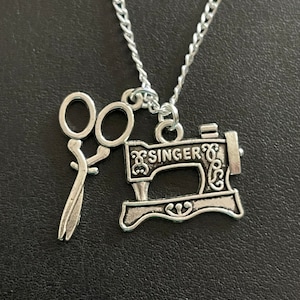 May include: Silver-toned necklace featuring a sewing machine charm with the word "SINGER" and a pair of scissors. The chain is a delicate silver color. The necklace is likely a craft or hobby-themed accessory.