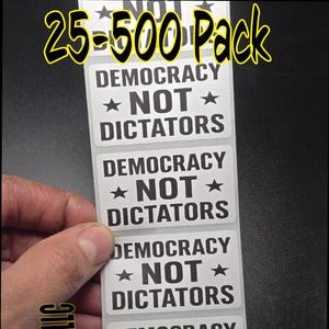May include: A pack of stickers with the text "DEMOCRACY ★ NOT ★ DICTATORS" in black on a white background. The text "25-500 Pack" is in yellow at the top. The brand name "SlickSticksLLC" is in yellow on the left side.