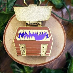 K&ouml;nnte beinhalten: Ein goldfarbener Metall-Schl&uuml;sselanh&auml;nger mit einem braunen Schatzkistendesign mit lila Zunge und wei&szlig;en Z&auml;hnen. Die Truhe hat goldfarbene Akzente und liegt auf einer Holzoberfl&auml;che.