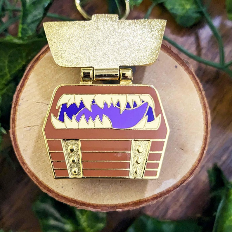 K&ouml;nnte beinhalten: Ein goldfarbener Metall-Schl&uuml;sselanh&auml;nger mit einem braunen Schatzkistendesign mit lila Zunge und wei&szlig;en Z&auml;hnen. Die Truhe hat goldfarbene Akzente und liegt auf einer Holzoberfl&auml;che.