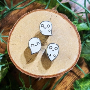 May include: Three white ghost pins with different facial expressions. The ghosts are on a light brown wooden surface.