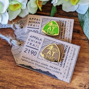 May include: Two gold-colored enamel pins with the Appalachian Trail logo. The pins are shaped like a triangle with the text "AT 2190" inside. The pins are on a brown paper tag with the text "Appalachian Trail" and "Leave No Trace" printed on it.
