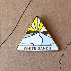 May include: Gold-colored enamel pin shaped like a triangle with a white sand dune and a yellow sun. The text "WHITE SANDS" is printed on the pin.