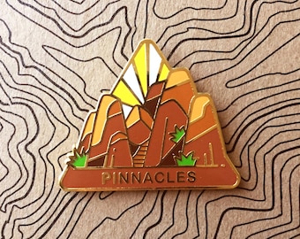 May include: A gold-colored enamel pin featuring a stylized illustration of a mountain range with a sunburst above. The mountain range is brown and orange with green trees. The text "PINNACLES" is printed in black on the bottom of the pin.