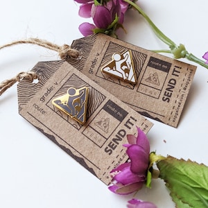 May include: Two gold-colored enamel pins with a climber silhouette on a mountain. The pins are on brown paper cards with the text "SEND IT!" and "grade: v" printed on them.