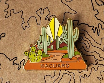 May include: A gold-tone enamel pin featuring a desert landscape with saguaro cacti, a sunset, and the word "SAGUARO" in the foreground.